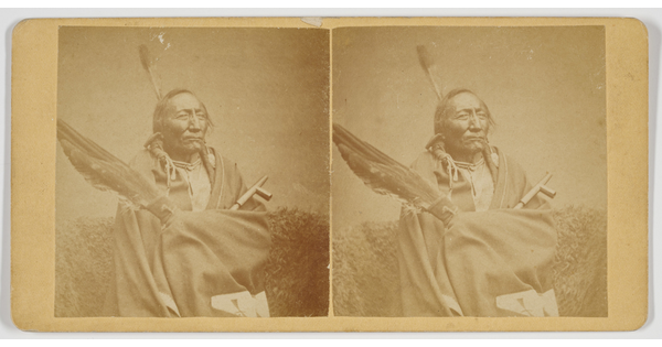 A sepia-toned stereograph of an Indigenous man holding a group of large feathers and a pipe.