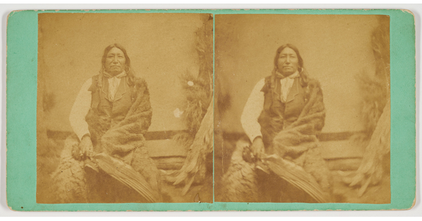 A sepia-toned stereograph of an Indigenous man wearing a vest and wrapped in a blanket, holding a feathers.