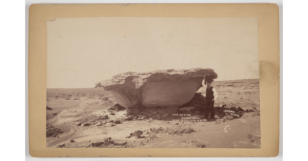 A sepia-toned photograph of a man standing next to a bowl-shaped rock formation in the desert.