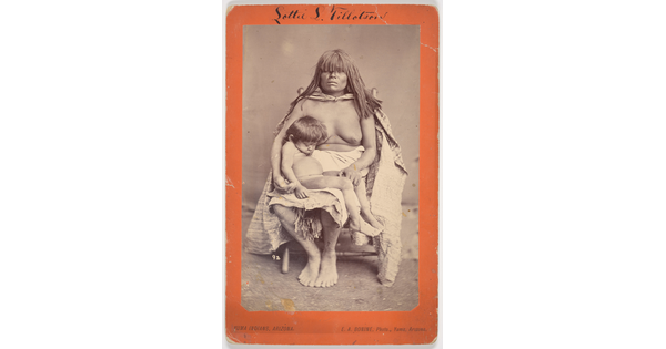 A black-and-white photograph of a seated Indigenous woman with long hair and bangs that almost cover her eyes, a blanket around her topless torso, and a sleeping child on her lap.