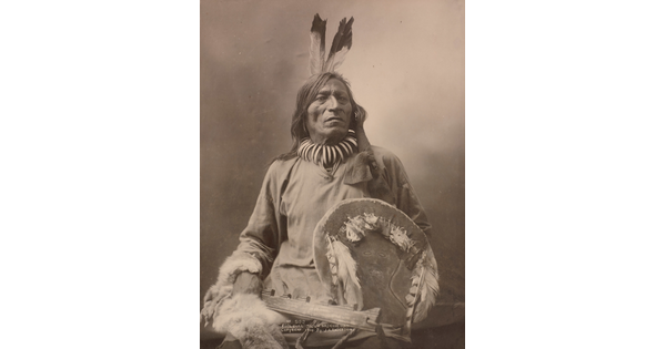 A black-and-white formal portrait photograph of a seated Indigenous man in traditional clothing holding a shield.