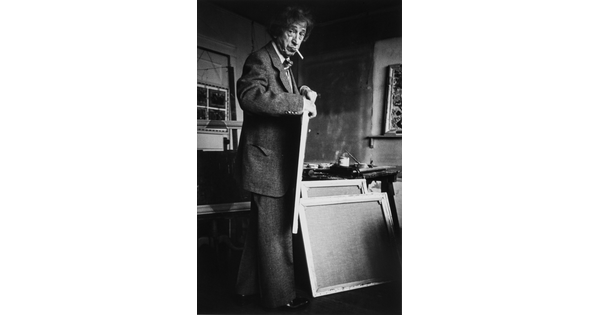 A black-and-white photograph of an older White man with a cigarette in his mouth holding a canvas in an artist's studio.