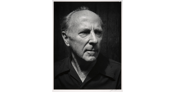 A black-and-white photograph of the head and shoulders of an older balding White man wearing a dark button-down shirt, head turned slightly.