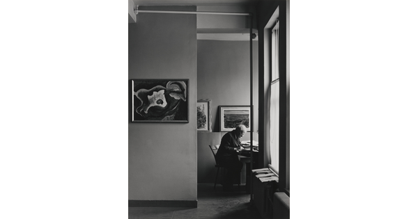 A black-and-white photograph of an older White man working at a desk in a room lit by sunlight through windows, artworks hanging on the walls.