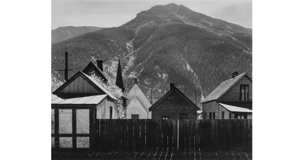 A black-and-white photograph of clapboard houses and a church at the base of a densely wooded mountain.