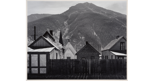 A black-and-white photograph of clapboard houses and a church behind a wood fence at the base of a densely wooded mountain.