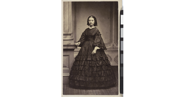 A black-and-white studio photograph of a White woman, dark hair styled in banana curls, wearing a long sleeved, full length, dark ruffled dress.