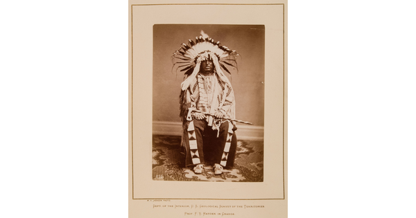 A black-and-white formal portrait photograph of a seated Indigenous man in full, traditional regalia with a headdress and tomahawk.