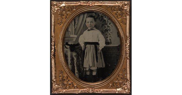 A black-and-white photograph in a decorative gold frame of a White child wearing a white shirt and plaid skirt, standing beside a table.