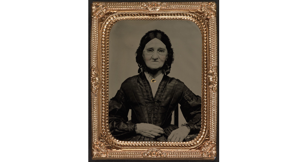A black-and-white photograph in a decorative gold frame of an elderly White woman in spectacles wearing a dark Victorian-style dress.