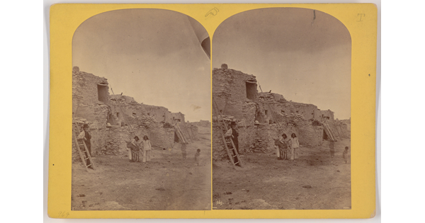Two almost identical side-by-side black-and-white photographs of several Indigenous people at a multi-level Native American pueblo.