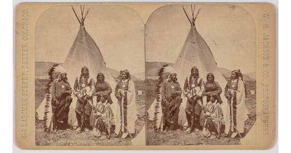 A black-and-white stereograph of five Indigenous people in traditional regalia standing in front of a tipi.