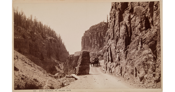 A sepia-toned photographic postcard of horses pulling a wagon on a dirt road between tall cliffs and rock formations.