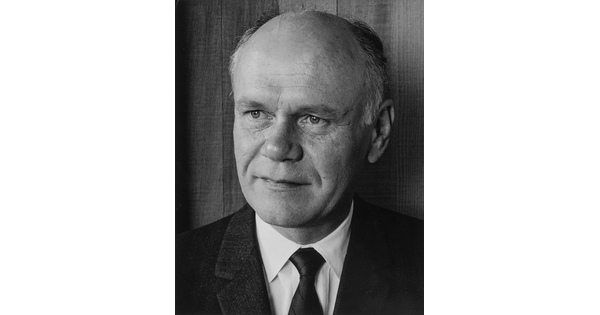 A black-and-white portrait photograph of a mostly bald White man gazing to the side wearing a dark jacket, dark tie, and white shirt.