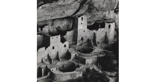 A black-and-white photograph of ruined stone buildings, some with towers, and several underground circular rooms all under the overhang of a cliff.