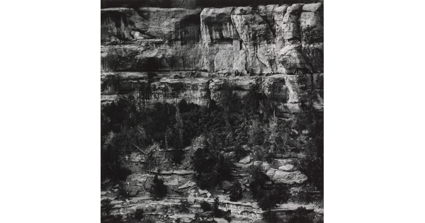 A black-and-white photograph of cliff dwellings in ruins on the side of a steep cliff.