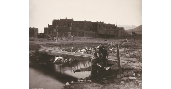 A black-and-white photograph of two Indigenous people by a footbridge over a creek with a large adobe pueblo in the background.