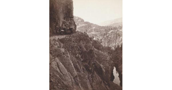 A sepia-toned photograph of a train travelling on the edge of a rocky, mountainside cliff.