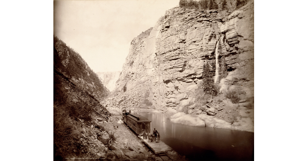 A black-and-white photograph of people on a train at the bottom of a canyon looking at a waterfall across a river.