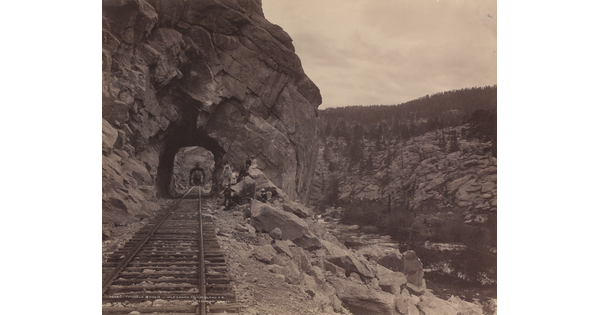 A black-and-white photograph of a train track stretching through a series of rock tunnels in the side of a mountain.