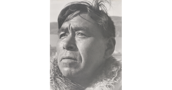 A black-and-white photograph of a close-up of a middle-aged Indigenous man with windblown hair and fur at his neck.