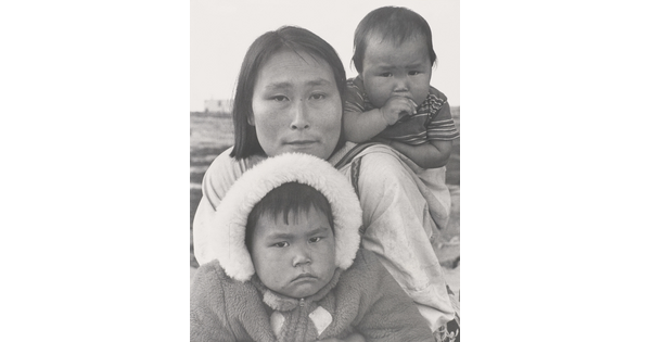 A black-and-white photograph of an Indigenous woman with a baby on her back and another child wearing a hooded coat in front of her.