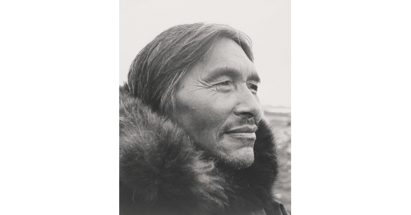 A black-and-white photograph of a close-up of a middle-aged Indigenous man, salt-and-pepper hair and wrinkles around his eyes, a mustache and a fur at his neck.