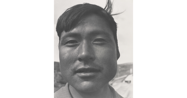 A black-and-white photograph of a close-up of an Indigenous man with short dark hair, one lock sticking up at back.