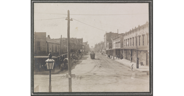 A black-and-white photograph of a western-style town with a trolley on tracks in the center of an unpaved street.