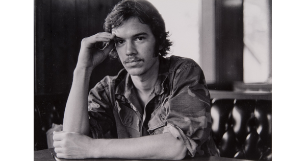A black-and-white photograph of young White man wearing a button-down shirt with the sleeves rolled up, one arm resting on the table in front of him, the other raised to his temple.