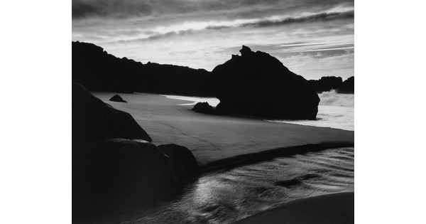 A black-and-white photograph of the sand and sea next to a shore covered with large rocks.