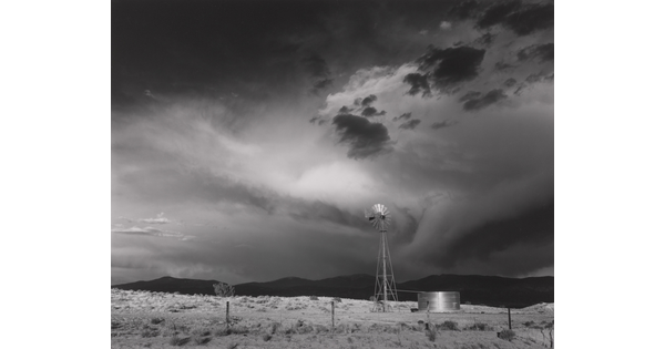 A black-and-white photograph of a windmill, black hills in the distance, under a stormy sky.