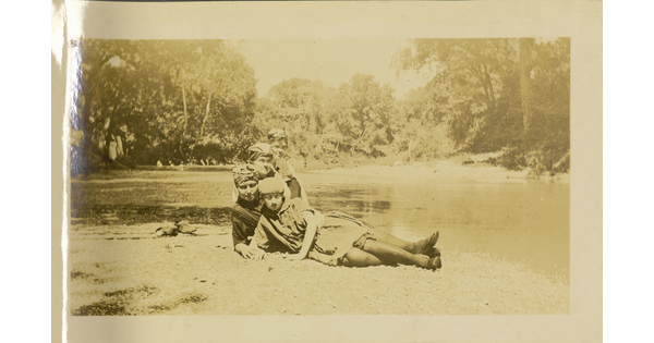 A faded sepia-toned photograph of four White women, all wearing headwraps, posing on the ground next to a river.