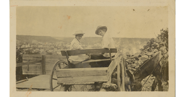 A sepia-toned photograph of two White women wearing sunhats sitting in a cart turned back to face the camera.