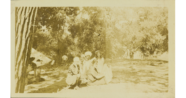 A faded sepia-toned photograph of six White women crouched next to a tent at a campsite.
