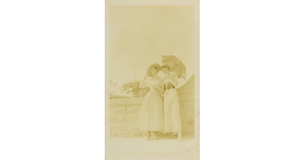 A faded sepia-toned photograph of two White women in sunhats and long skirts, one holding an open parasol, standing next to a stone wall.