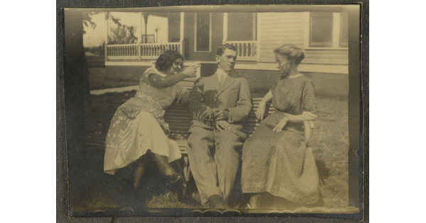 A sepia-toned photograph of two White women and a White man seated between them on a bench in front of a large house.