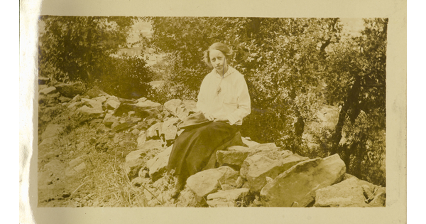 A sepia-toned photograph of a White woman wearing a light blouse and long dark skirt sitting on rocks holding a canvas hat in her lap.