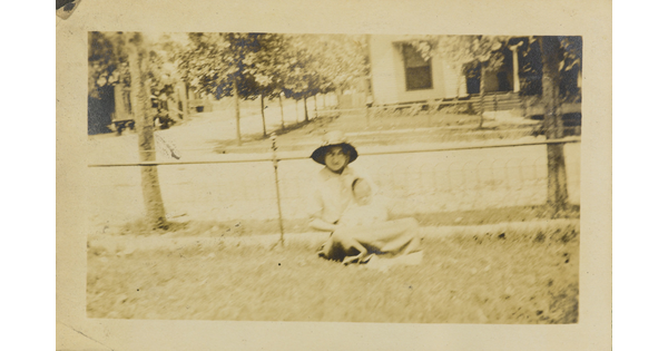 A faded sepia-toned photograph of a White woman in a sunhat holding a baby wearing a bonnet on her lap sitting on grass in front of a fence.