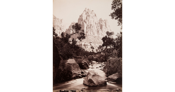 A black-and-white photograph of a stream surrounded by boulders and vegetation and a rugged cliff in the background.
