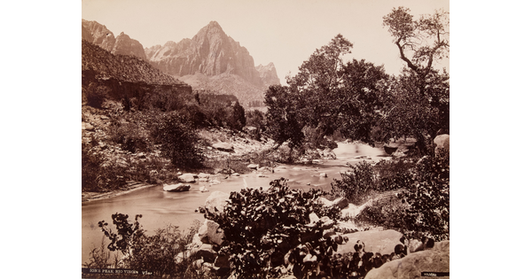 A black-and-white photograph of a rocky river surrounded by vegetation, and a tall mountain rising in the background.