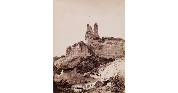 A sepia-toned photograph of a tall rock formation on top of large boulders above scrubby vegetation.