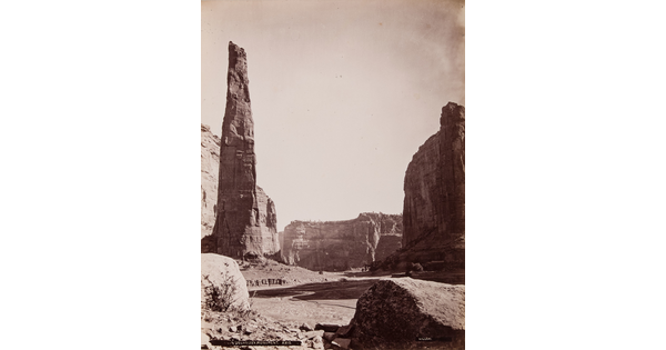 A sepia-toned photograph of a tall, narrow rock formation opposite a butte with a horizontal rock formation in the background.