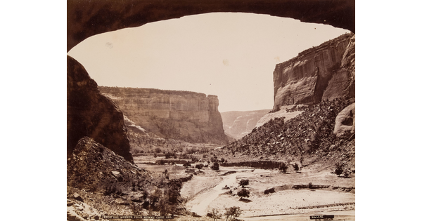 A sepia-toned photograph of tall cliffs on either side of a dry gully, framed by the bottom of an arch.