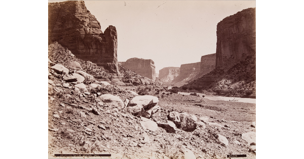 A sepia-toned photograph of buttes and other rock formations rising above a narrow canyon covered with loose rocks.