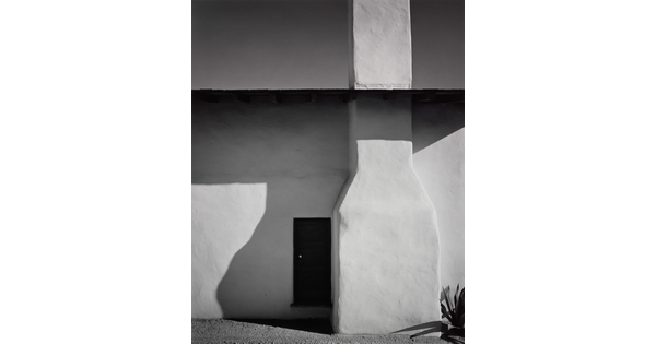 A black-and-white photograph of the side of an adobe house with the foundation of a chimney, shadows on the wall, and a door.