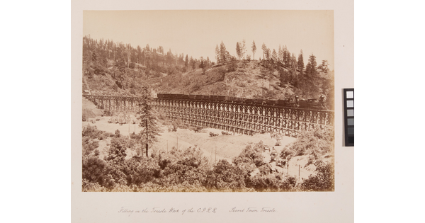 A sepia-toned photograph of a train travelling across a mountain valley with onlookers at base of the trestle.