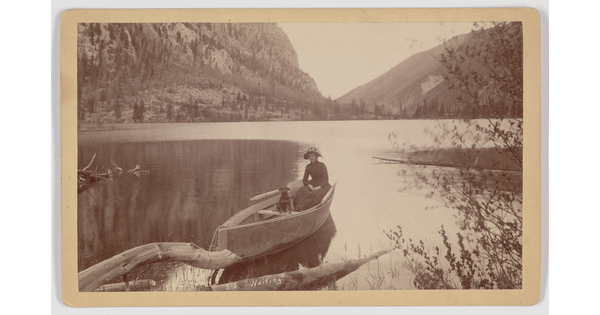 A sepia-toned photograph of a woman and a dog in a canoe on a still lake in the mountains.
