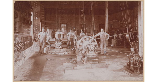 A sepia-toned photograph of several White men posing by industrial equipment and gears in a brick building.