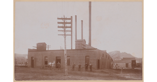 A sepia-toned photograph of men standing in front of a one-story brick building with smokestacks.
